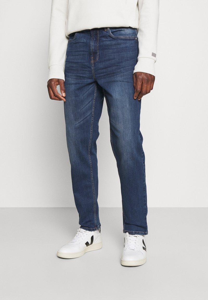 Dark blue denim jeans with a straight fit. Features five-pocket design, subtle stitching, and worn-in texture. White sneakers complete the look.