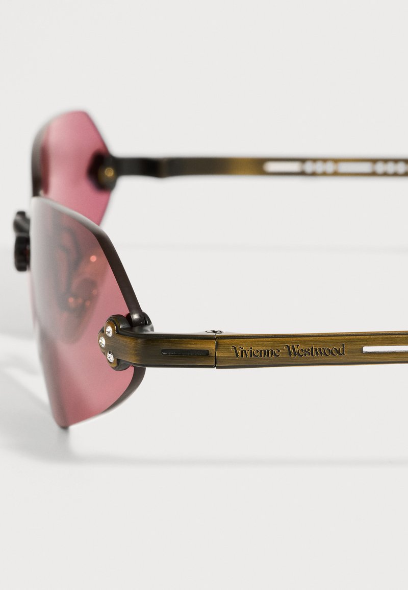 Lunettes de soleil avec verres roses et branches métalliques bronze, décorées de petits strass près des charnières et gravées de la marque Vivienne Westwood.