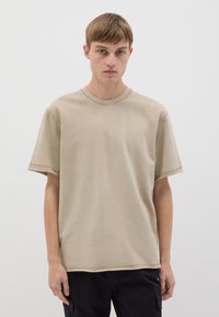 T-shirt in cotone beige con collo rotondo, maniche corte e dettagli di cucitura visibili, abbinato a pantaloni scuri, su uno sfondo bianco.