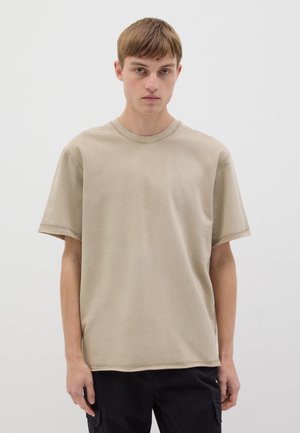 Camiseta de algodón beige con cuello redondo, mangas cortas y detalles de costura visibles, combinada con pantalones oscuros, sobre un fondo blanco.