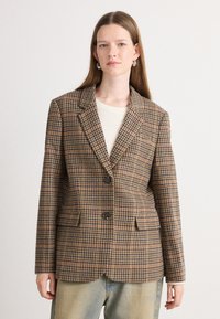Blazer à carreaux marron avec revers cranté, deux poches avant et deux boutons ; associé à un haut crème et un jean bleu clair.