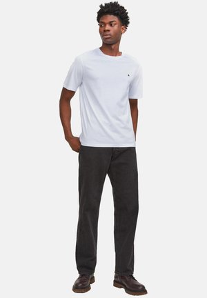 Homme debout les mains dans les poches, portant un T-shirt blanc uni à manches courtes, un jean noir et des chaussures marron, regardant vers sa gauche.