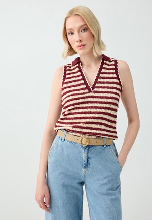 Blonde vrouw draagt een mouwloos rood-wit gestreept gebreid topje met een kraag en lichtblauwe hooggetailleerde jeans met een beige riem, staand tegen een witte achtergrond.