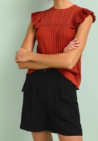 Top sin mangas de color óxido con canalé, mangas acampanadas y detalles de encaje, combinado con pantalones cortos negros con bolsillos laterales y un ajuste entallado.