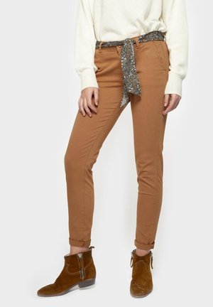 Vrouw draagt beige slim-fit broek met een bloemenstoffen riem, een witte trui en bruine suède enkelhoge laarzen met zijritsen.