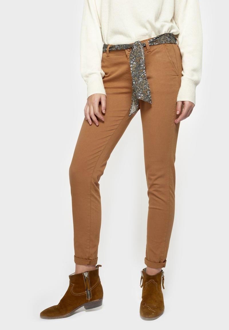 Femme portant un pantalon slim beige avec une ceinture en tissu floral, un pull blanc et des bottines en suède marron avec des fermetures éclair sur le côté.