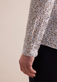 Langärmelig overhemd met een lichtblauwe basis, versierd met een klein bloemenpatroon in bruin en groen. Knopenmanchetten met een gladde textuur.