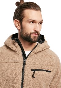 Chaqueta de forro polar beige con capucha, con cierre frontal y bolsillo, detalles en negro y tela texturizada.