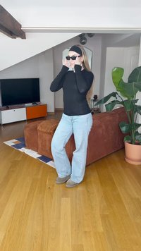 Fekete hosszúujjú felső, világoskék bővülő farmernadrág, fekete napszemüveg és bézs lábbeli, egy modern szobában állva, ahol egy növény és egy kanapé is található.