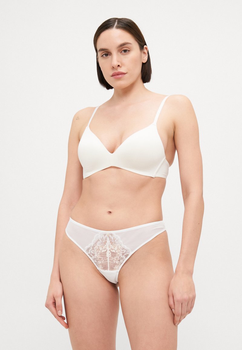 Soutien-gorge blanc en tissu lisse avec des bretelles fines; string blanc assorti avec un design en dentelle à l'avant. Le modèle se tient devant un fond uni.