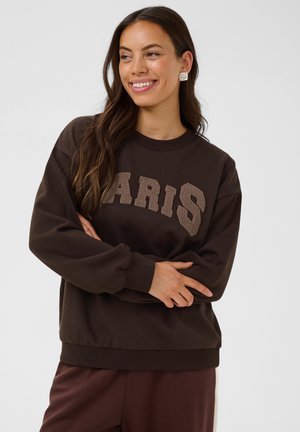 Femme avec de longs cheveux foncés portant un sweat-shirt marron avec le texte texturé "PARIS", souriante et les bras croisés devant un fond uni.