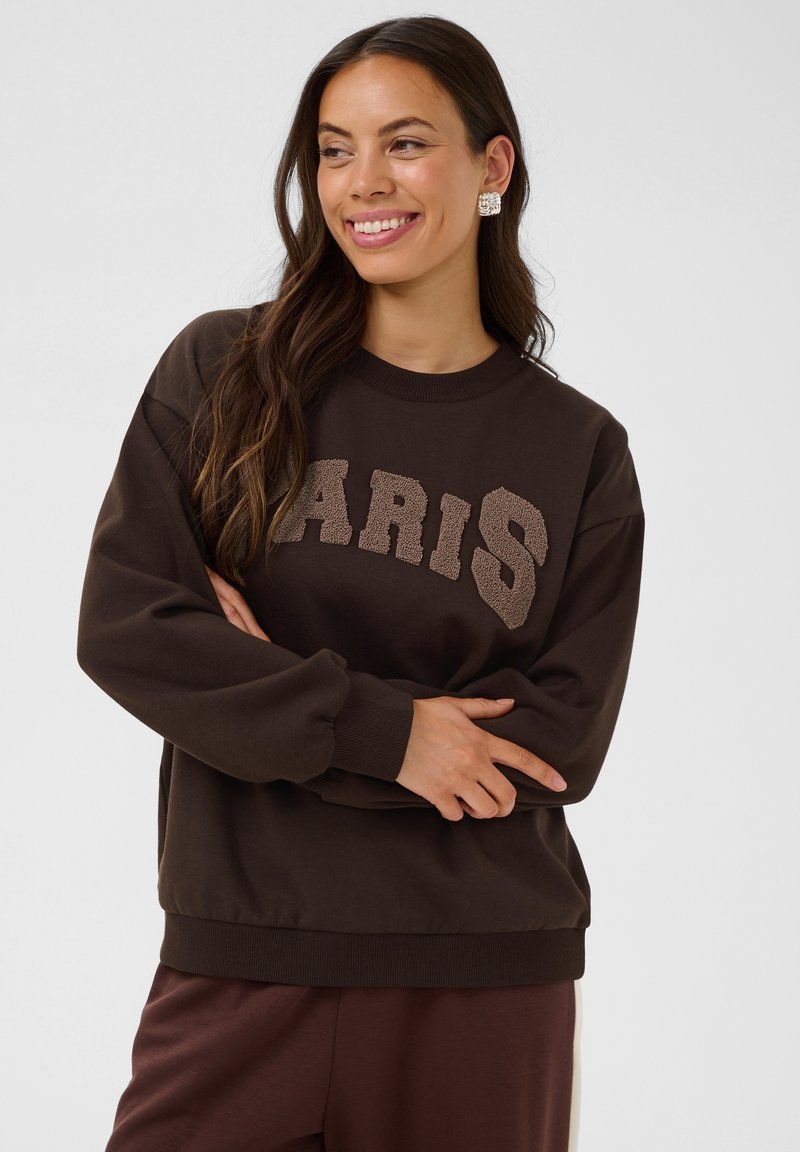Femme avec de longs cheveux foncés portant un sweat-shirt marron avec le texte texturé "PARIS", souriante et les bras croisés devant un fond uni.