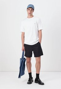 White short-sleeve t-shirt, black shorts, black socks, and black shoes. Holds a blue denim tote bag. Light blue cap. Minimalist design.