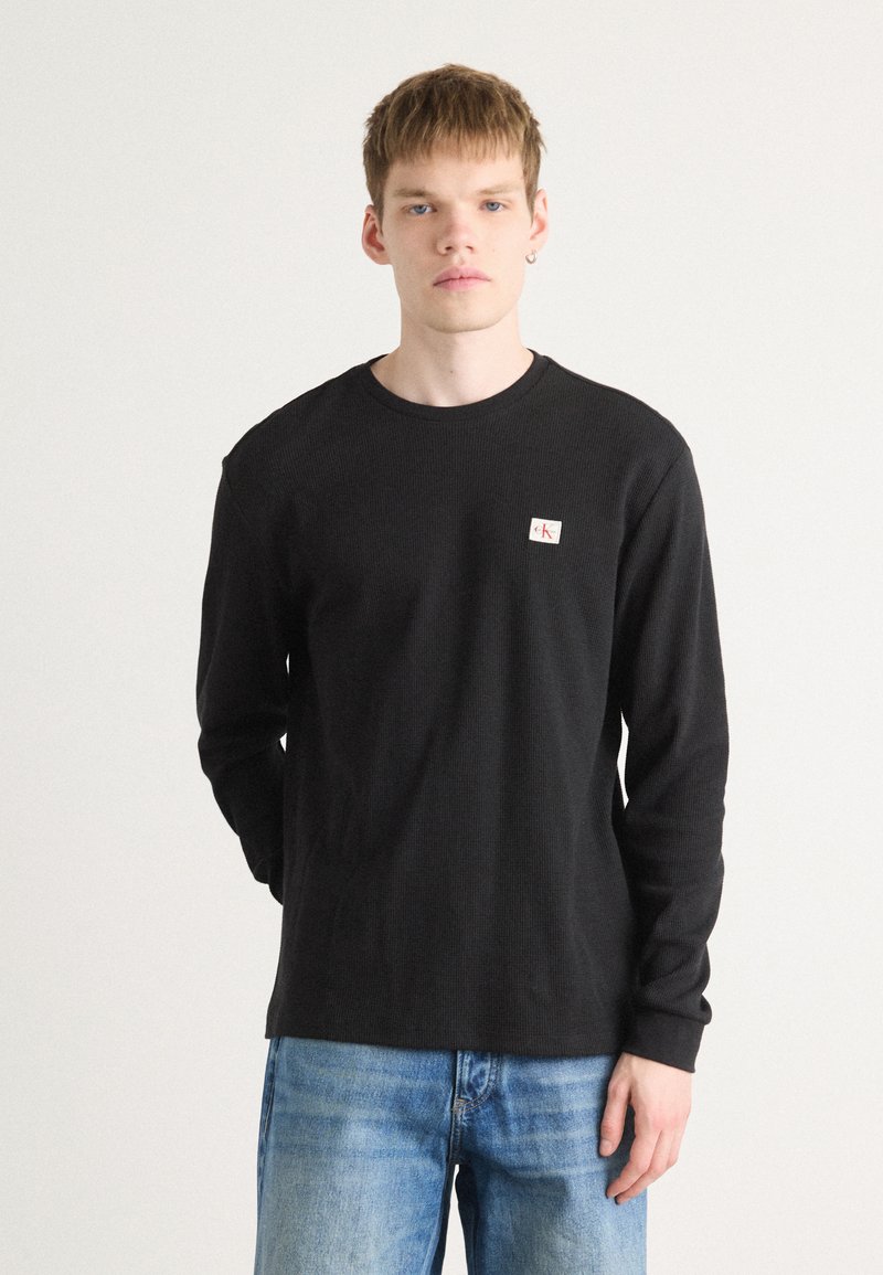 Zwarte langemouwen shirt van een textuurstof met een ronde halslijn, voorzien van een klein logo-opdruk op de linkerborst. Gegdraag in combinatie met blauwe spijkerbroeken.