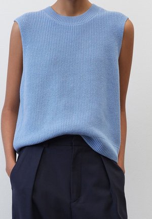 Lichtblauw, mouwloze gebreide top met een geribbelde textuur en een ronde halslijn, gecombineerd met donkere broeken, weergegeven tegen een effen achtergrond.