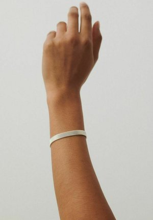 Ruka smeđe puti podignuta na jednostavnoj svijetloj pozadini, na zapešću nosi tanku narukvicu srebrne mrežaste teksture.
