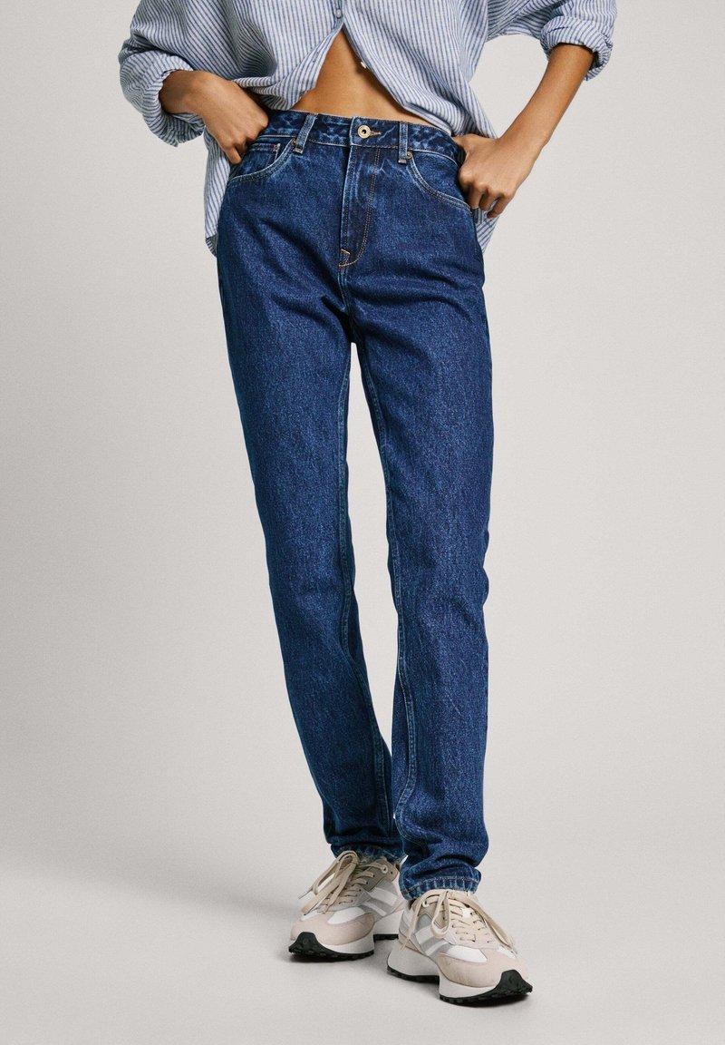 Personne portant un jean taille haute bleu, des baskets gris clair et beige, et une chemise rayée bleue partiellement rentrée.
