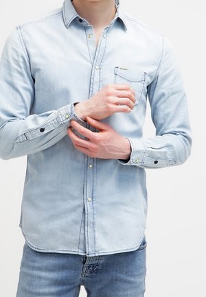 Homme ajustant le poignet d'une chemise en jean bleu clair, portant un jean bleu sur un fond blanc uni.