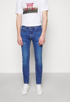 Person trägt blaue Slim-Fit-Jeans, weiße Sneaker und ein weißes T-Shirt mit einem roten Torii-Tor und dem Schriftzug „HUGO“.