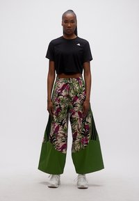 Mujer de pie mirando hacia adelante, vestida con un top negro corto y pantalones de pierna ancha con estampados florales verdes y rosas, y de pájaros.