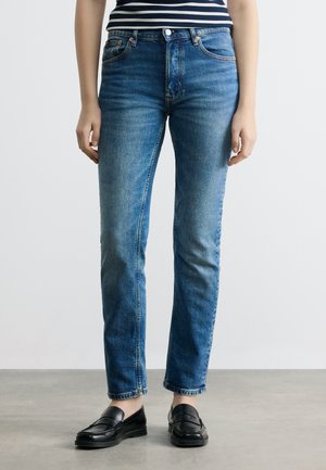 Person iført blå straight-leg jeans med mellemhøjt talje og sorte loafers, stående på en grå gulv mod en hvid baggrund.