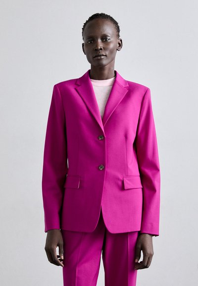 Blazer fucsia su misura con due bottoni e tasche a bavero, realizzato in tessuto liscio, abbinato a una maglietta rosa chiaro e pantaloni coordinati.