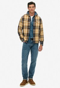 Geruite flanellen overhemden in oranje, geel en marineblauw, gedragen over een denimoverhemd, gecombineerd met blauwe jeans en bruine laarzen. Bevat knoopdetails.