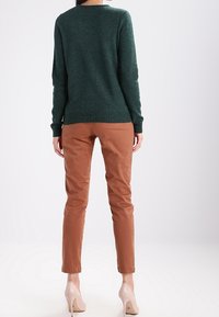 Grüner Strickpullover mit rundem Ausschnitt, kombiniert mit beige-farbenen, schmal geschnittenen Hosen. Der Pullover hat gerippte Bündchen und Saum; glatte Textur, neutrale Schuhe.