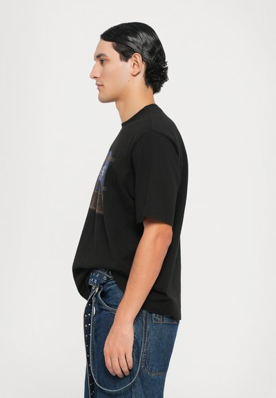 Camiseta de algodón negra con mangas cortas, corte relajado, que presenta un estampado gráfico en la parte frontal. Llevada con jeans de mezclilla azules y hardware decorativo.