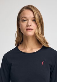 Granatowa koszula z długim rękawem wykonana z miękkiego materiału, z okrągłym dekoltem i małym czerwonym haftowanym logo golfisty na lewej piersi.