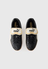 Zwart-witte voetbalschoenen met een opvallende klittenbandsluiting, synthetisch materiaal, getextureerde details en rubberen zolen met een klein logo.