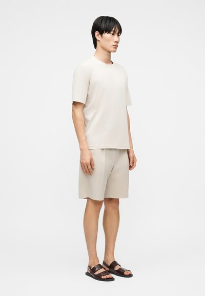 Άνδρας που στέκεται φορώντας μπεζ πουκάμισο με κοντά μανίκια και αντίστοιχο σορτς με σκούρα καφέ ανοιχτά σανδάλια σε λευκό φόντο.