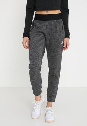 Kvinde iført mørkegrå Adidas træningsbukser med lynlåslommer, sort cropped langærmet top og hvide sportssko, stående indendørs.