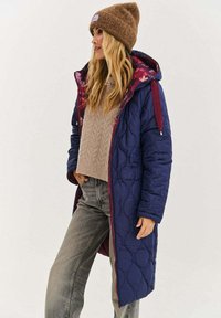 Granatowy pikowany długi płaszcz z bordowymi akcentami, kwiecistą podszewką i kapturem ze ściągaczem, zestawiony z lekkim swetrem i szarymi dżinsami.