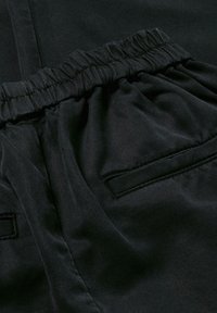 Zwarte stoffen pantalon met een elastische tailleband, voorzien van zijzakken en een gladde textuur. Stiksels zijn zichtbaar op de zakken.