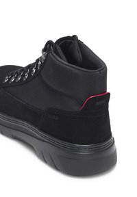 Zapatilla deportiva de caña alta de ante negro con suela de goma, ojales metálicos, cuello acolchado y un detalle en rojo en el talón.