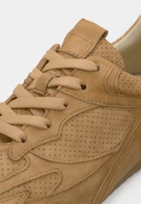 Τακούνι από καφέ σουέτ με διάτρητα πάνελ, επίπεδα κορδόνια και επένδυση στο γιακά. Η σόλα έχει ελαφρά ανύψωση και υφή για καλύτερη πρόσφυση.