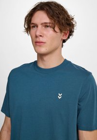 Kortärmad T-shirt med rund hals i teal bomullstyg, med en liten vit chevron-logotyp på vänster bröst. Slät textur, avslappnad design.