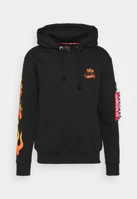 Sweat à capuche noir avec poche avant, logos Alpha Industries et Hot Wheels orange sur la poitrine, motif de flammes sur la manche gauche, et étiquette rouge "Remove Before Flight" sur le bras droit.