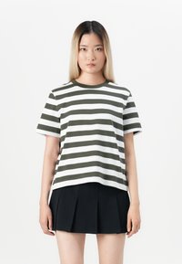 Ung kvinna med långt rakt hår, iförd en olivgrön och vit randig T-shirt samt en svart plisserad kjol, som står mot en vit bakgrund.