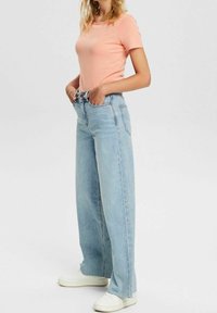 Hellrosa, kurzärmliges T-Shirt kombiniert mit hochgeschnittenen, weit geschnittenen, hellblauen Jeans und weißen Turnschuhen. Glatter Stoff und klassischer Schnitt.