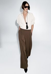 Bílá volná košile, hnědé vysoké kalhoty a černé sluneční brýle. Černý náhrdelník s třásněmi jako akcent. Hladká, strukturovaná látka.
