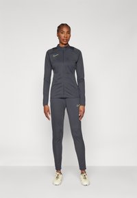 Woman standing in dark gray Nike tracksuit with neon logos and white soccer cleats against plain white background.