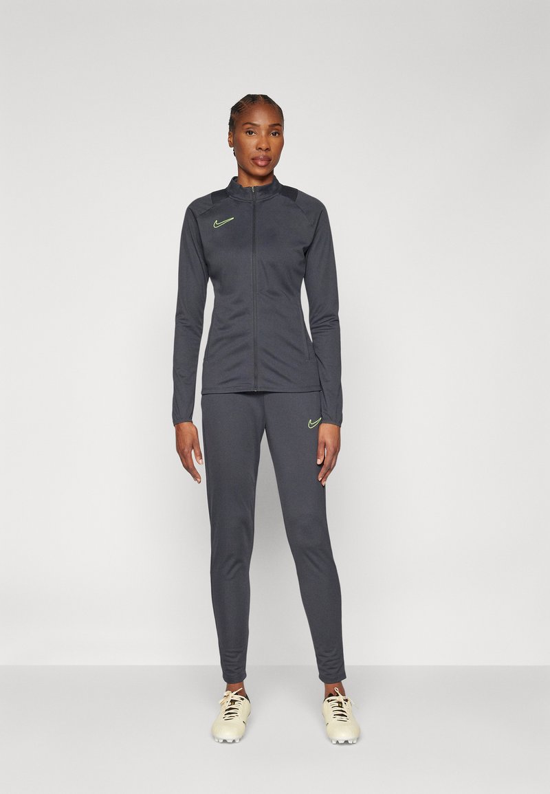 Woman standing in dark gray Nike tracksuit with neon logos and white soccer cleats against plain white background.