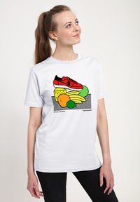 Bílý tričko s barevným grafikou červeného tenisky a různými plody, včetně banánů a pomerančů, na pruhovaném pozadí.