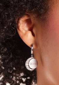 Zilveren creolen met een ronde hanger waarop "GUESS" is gegraveerd, versierd met heldere rhinestones, tentoongesteld op textuurachtig, donker haar.