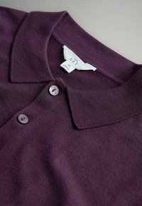 Ljubičasta polo majica s klasičnim ovratnikom, dvostrukim gumbom i rebrastom teksturom. Na etiketi su informacije o brendu i veličini.