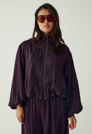 Femme aux longs cheveux foncés portant de grandes lunettes de soleil teintées rouges et une veste zippée violet foncé à manches larges sur un fond uni.