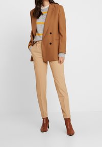 Blazer marron à double boutonnage sur un pull gris à rayures jaunes, associé à un pantalon beige ajusté et des bottines brunes.