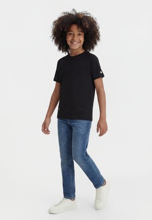 Smilende barn med krøllete hår som har på seg en svart t-skjorte, blå jeans og hvite joggesko, står foran en ensfarget hvit bakgrunn.